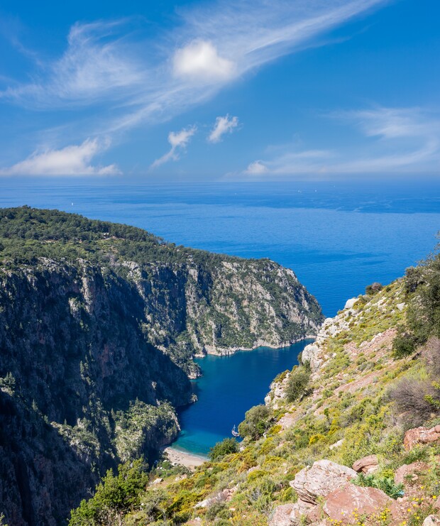 A cliff overlooks a deep blue cove with clear skies above. The surrounding landscape is lush with vegetation, and the sea stretches out to the horizon.